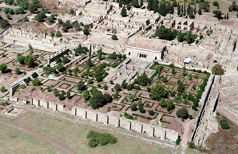 An aerial view of trees and rectilinear hedges, behind which are architectural ruins.