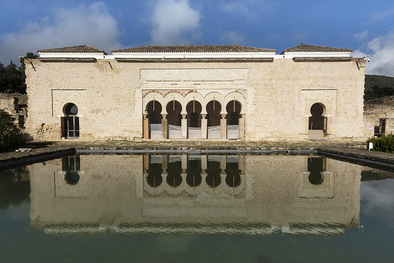 A rectangular façade pierced by five horseshoe arches at the center and one at either end. Topped by a tile roof and facing a rectangular pool of water.