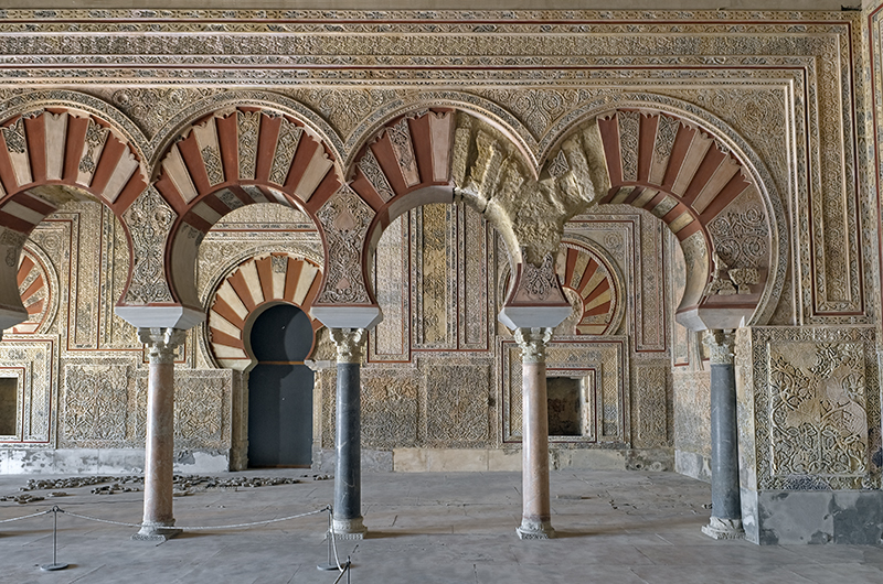 Four horseshoe arches supporting an entablature decorated with vegetal, geometric, and epigraphic motifs. The view leads to a further wall with similar arches and decoration.