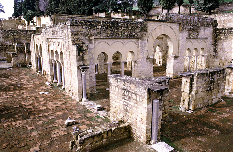 The oblique view of a stone wall facing a rectangular stone structure with horseshoe arches and set on brick.