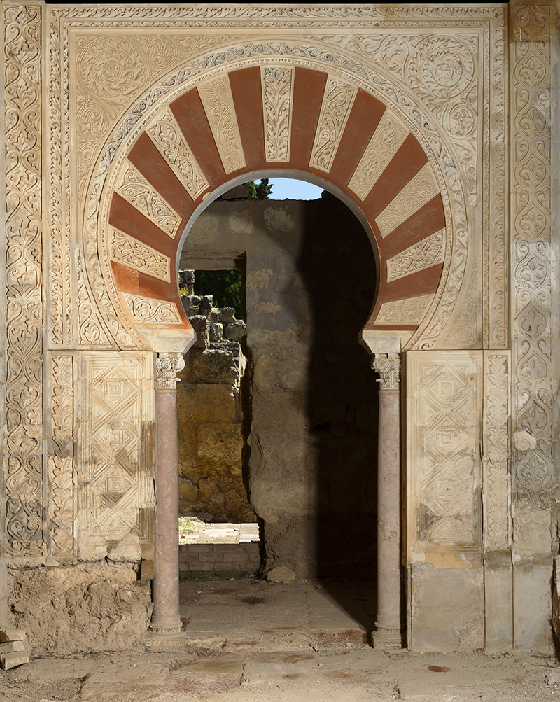 A horseshoe arch set into a wall with geometric and vegetal carved designs, and a view into further rooms.